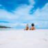 two women lying on white sand facing beach under blue sky